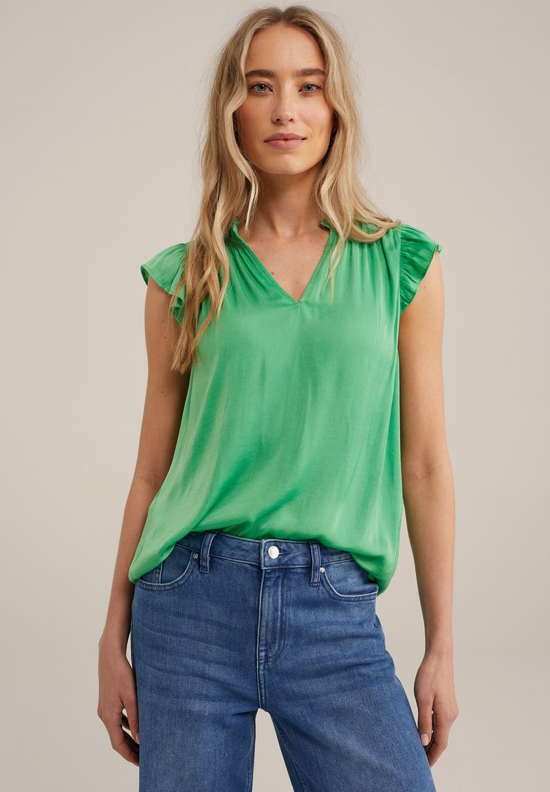 Blusa verde con scollo a V e maniche corte a volant, realizzata in un tessuto morbido. Indossata sopra jeans blu con vestibilità classica e dettagli minimi.