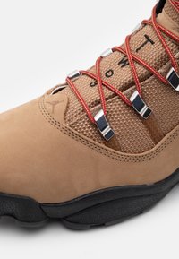 Gros plan sur une basket beige avec des lacets rouges, une semelle noire et une languette en tissu texturé embossée du mot « TWO » et d'un petit logo Jumpman.