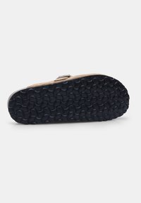 A talpája egy textúrázott fekete gumipattern-t tartalmaz, körkörös fogantyúkkal, kiegészítve egy világos bézs felsőrész és fém csat részlet.