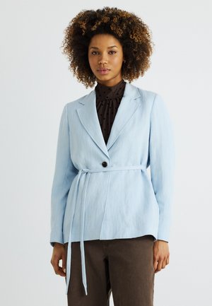 Femme aux cheveux bouclés portant un blazer bleu clair ceinturé sur un chemisier noir à pois et un pantalon foncé, debout devant un fond blanc.