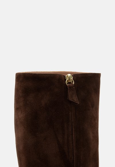 Partie supérieure d'une botte en daim marron foncé avec une fermeture éclair dorée visible et une languette assortie en daim sur un fond gris clair.