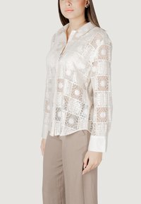 Valgest pitspluus, millel on nööpidega ees, pikad varrukad ja mustriline disain, mis sisaldab lillemotiive. Kerge kangas ja kaarjas kaelus.