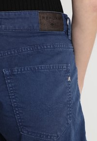 Jean en denim bleu avec un patch en cuir marron portant l'inscription "REPLAY". Dispose d'une poche arrière avec un motif de couture subtil et du matériel argenté.