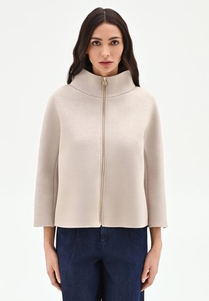 Femme aux cheveux longs et foncés portant une veste beige à fermeture éclair avec un col montant et des manches trois-quarts, associée à un pantalon bleu foncé.