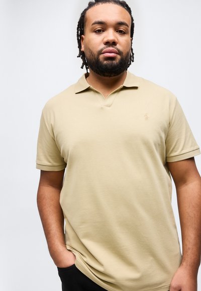 Άνδρας με πλεξούδες στα μαλλιά και γενειάδα, φορώντας μπεζ πόλο με τα χέρια στις τσέπες, μπροστά σε απλό λευκό φόντο.