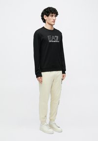 Fekete pulóver "EA7 Emporio Armani" logóval, kerek nyakkal és hosszú ujjakkal, bézs melegítőnadrággal és fehér sportcipővel párosítva.