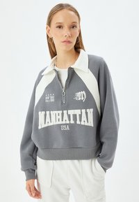 Grå cropped sweatshirt med hvide accenter, høj krave og halv lynlås. Har "MANHATTAN USA" trykt i hvidt samt dekorative elementer.
