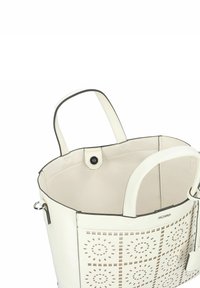 White leather tote bag with cut-out floral pattern, dual handles, and a smooth finish. Interior is beige, with a snap button closure.