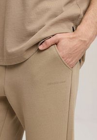 Beżowa bawełniana koszula o luźnym kroju, połączona z dopasowanymi joggerami z kieszeniami po bokach oraz wytłoczonym logo.