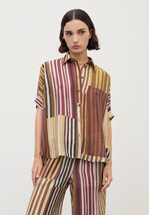 Donna che indossa una camicia ampia a righe multicolori con bottoni e pantaloni ampi abbinati su uno sfondo bianco uniforme.