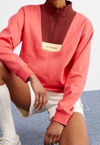 Felpa con zip color corallo e marrone, caratterizzata da un pannello a contrasto con una toppa logo, texture morbida e vestibilità rilassata. Abbinata a pantaloni corti color crema.