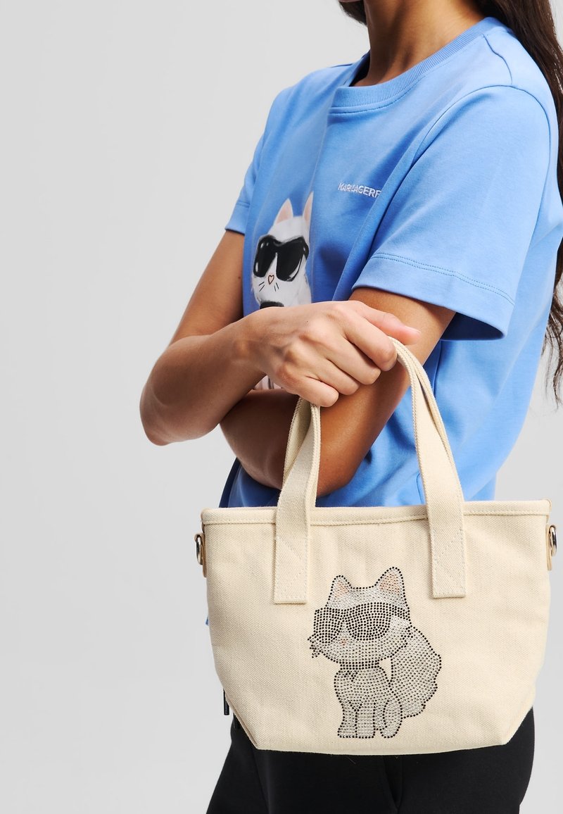 Personne en chemise bleue avec un motif de chat portant des lunettes de soleil tient un sac cabas beige présentant un motif similaire de chat avec lunettes de soleil.