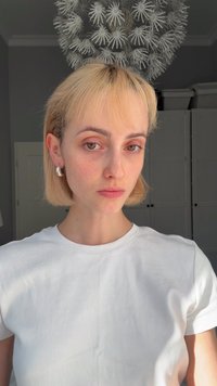 T-shirt en coton blanc porté par une personne aux cheveux blonds clairs et aux boucles d'oreilles anneaux, présentant une texture simple et de légères taches de rousseur naturelles sur le visage.