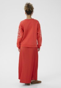 Femme avec des cheveux bouclés portant un sweat à capuche rouge avec des designs floraux blancs sur les manches et une jupe plissée rouge assortie, chaussée de baskets marron.