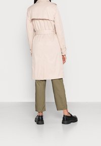 Beige trenchcoat med midjebälte, krage med slag och uppvikta detaljer. Matchas med olivgröna byxor och svarta robusta skor.