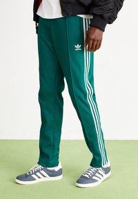 Zelené sportovní kalhoty z elastické látky, s třemi bílými pruhy po stranách, logem Adidas a zúženým střihem. Doplněné šedými teniskami.
