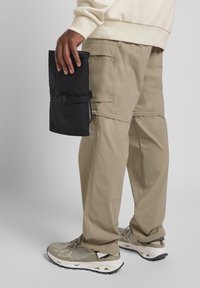 Borsa nera a rotolo tenuta in mano, indossata con pantaloni cargo beige. Scarpe grigie con design a rete e suole bianche. Aspetto semplice e funzionale.