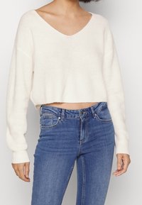 Pull court en maille crème avec un col en V et des manches longues légèrement oversize, associé à un jean bleu taille haute.