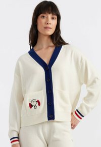 Cremfarbener Cardigan mit marineblauem Kragen und Knöpfen, der ein Snoopy-Graphic auf der linken Tasche und gestreifte Akzente an den Ärmeln aufweist.
