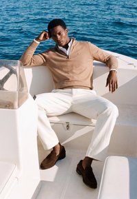 Homme portant un pull beige et un pantalon blanc, assis détendu sur un bateau blanc avec de l'eau bleue en arrière-plan.