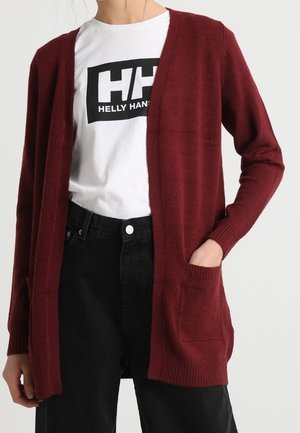Person iført en hvid Helly Hansen logo t-shirt, løs vinrød åben cardigan med lommer og høj talje sorte bukser.