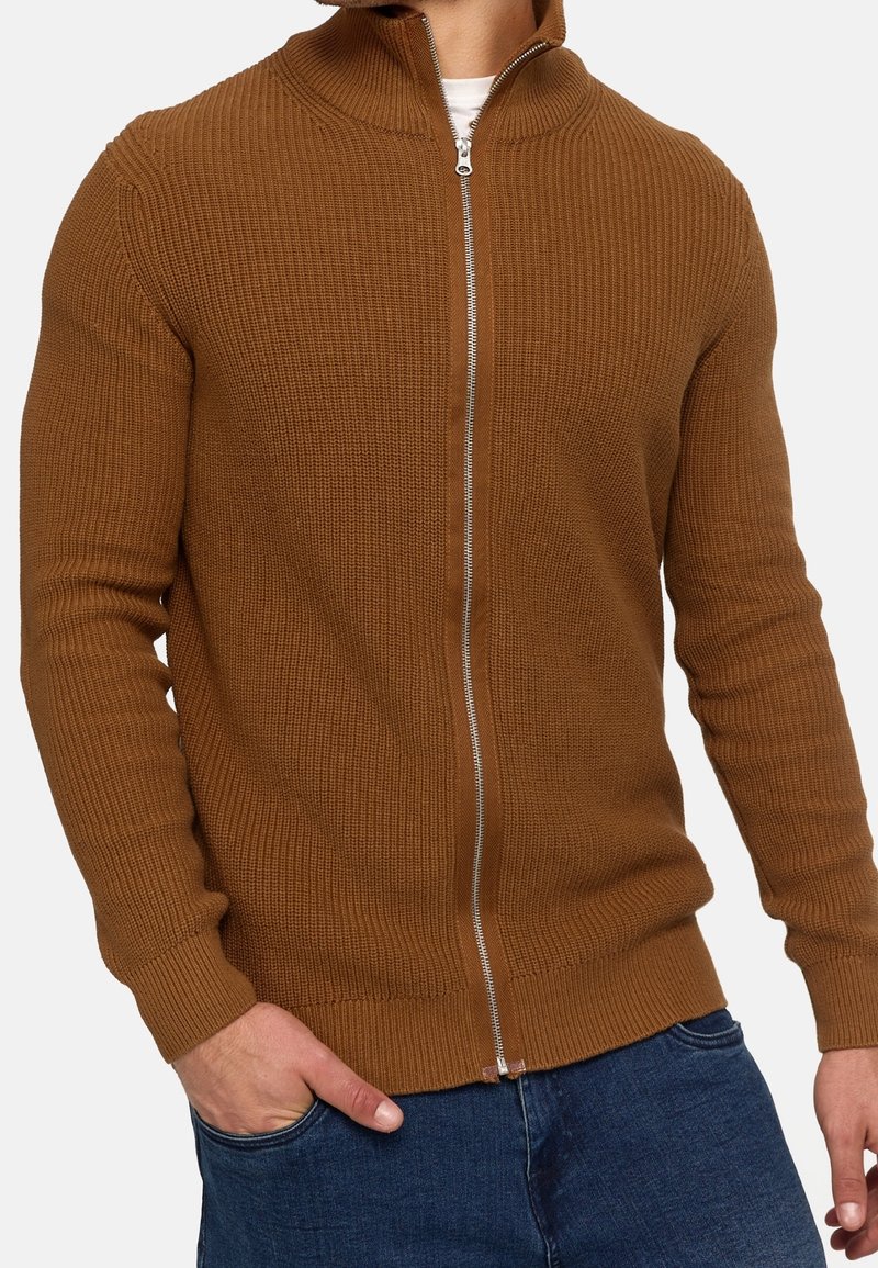 Homme portant un pull en maille marron à fermeture éclair sur une chemise blanche, avec une main dans la poche d'un jean bleu, sur fond blanc.