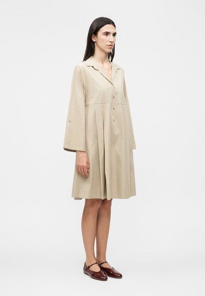 Γυναίκα με μακριά σκούρα μαλλιά φοράει μια άνετη μπεζ φόρεμα μέχρι το γόνατο με μακριά μανίκια και κουμπιά, συνδυασμένη με σκούρα κόκκινα παπούτσια Mary Jane.