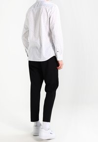 Vit långärmad skjorta med små flerfärgade mönster, kombinerad med svarta smalna byxor och vita sneakers, sedd bakifrån.