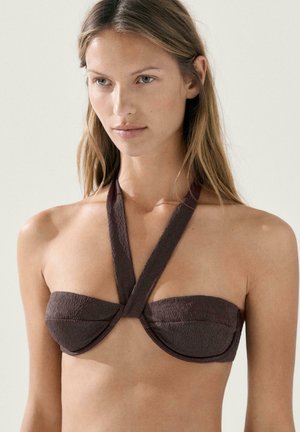 Woman with long blonde hair wearing a textured dark brown halter bikini top against a plain light background.