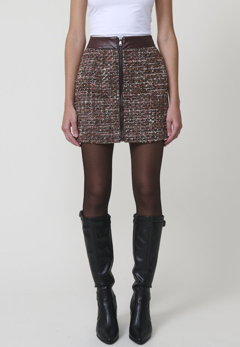 Minifalda de tweed en marrón, rojo y blanco con cremallera frontal y cinturilla de cuero sintético marrón; combinada con botas negras hasta la rodilla.