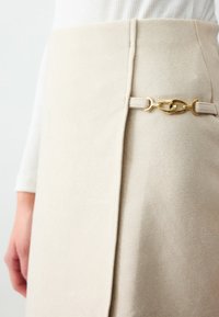 Beige rok met een zachte textuur, voorzien van een gouden kettingaccent en een zijkantwikkeling, die moderne en minimalistische styling benadrukt.