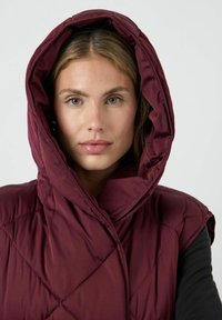 Burgunderröd pufferväst med stor huva, diamond-quiltad textur och ärmlös design. Slät tyg med en mjuk lyster.