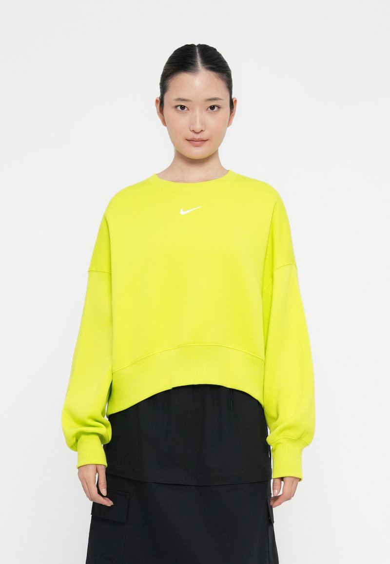 Neon rumena prevelika bombažna majica s ribastimi manšeti in robom, ki ima majhen bel Nike logo na prsih. Nosi se preko črne krila.