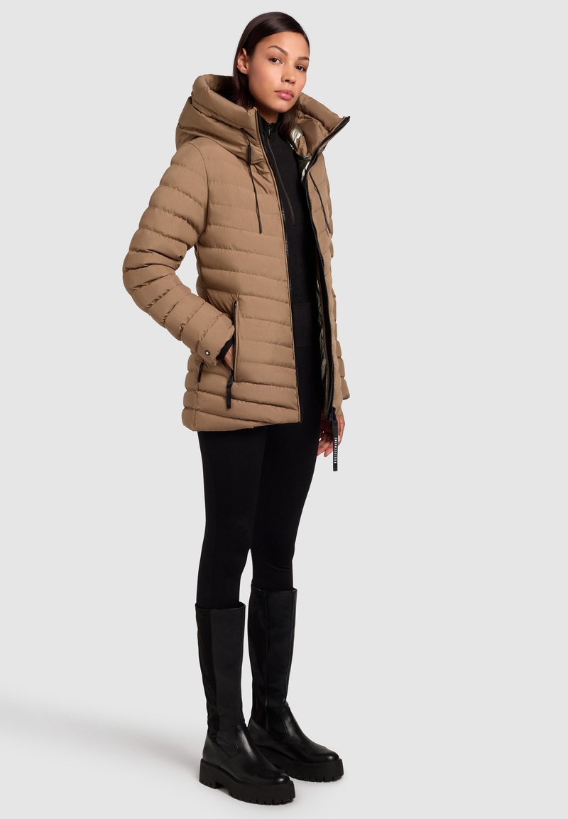 Beige gefütterte Jacke mit hohem Kragen, Frontreißverschluss und seitlichen Taschen. Getragen über einem schwarzen Oberteil und kombiniert mit schwarzen kniehohen Stiefeln.