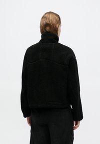 Jachetă din fleece neagră, cu guler înalt și o suprafață texturată, moale. Prezintă un design scurt și cusături pe spate.