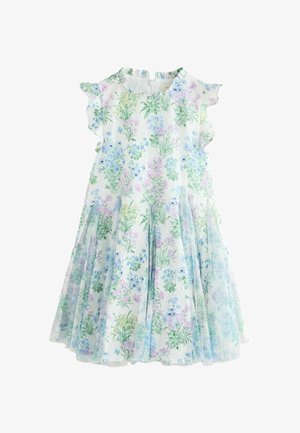 Fiatal lány göndör hajjal, pasztell virágos ruhában és bézs csillogó balettcipőben, semleges háttér előtt állva.