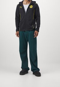 Uomo in giacca nera con zip e logo giallo del Borussia Dortmund, pantaloni turchesi, scarpe nere e maglietta bianca, in piedi su uno sfondo neutro.