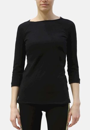 Vrouw die een zwart nauwsluitend shirt met lange mouwen en een zwarte broek met beige decoratieve zijkanten draagt, staand tegen een effen achtergrond.