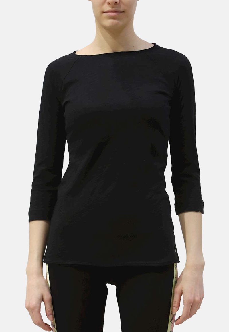 Femme portant un haut noir ajusté à manches longues et un pantalon noir avec des panneaux décoratifs latéraux beiges, debout devant un fond uni.