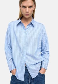 Jasnoniebieska, pasiasta koszula z długim rękawem, z kołnierzykiem zapinanym na guziki, w luźnym kroju i z miękkiego materiału; ma delikatne pionowe paski i zaokrąglone mankiety.