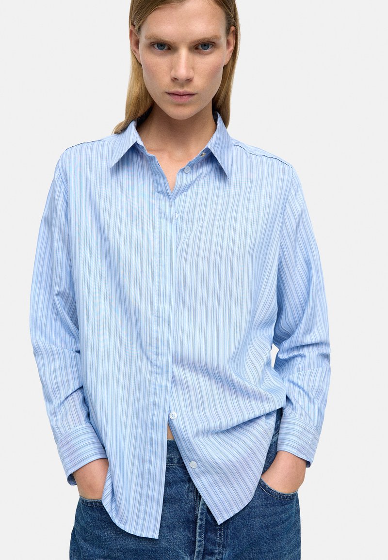 Jasnoniebieska, pasiasta koszula z długim rękawem, z kołnierzykiem zapinanym na guziki, w luźnym kroju i z miękkiego materiału; ma delikatne pionowe paski i zaokrąglone mankiety.