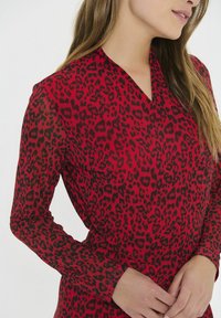 Hnedá blúzka s leopardím vzorom, dlhé rukávy, V-krk a priesvitný materiál, s kontrastnými čiernymi leopardími škvrnami a priliehavým strihom.