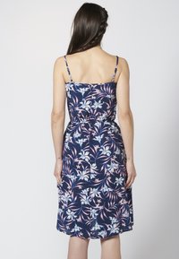 Laivastonsininen kukkamekko, jossa on vartalonmyötäinen vyötärö, ohuet säädettävät olkaimet ja polvea hipova helma. Mekossa on pinkkejä ja sinisiä kasvi-aiheisia kuvioita.