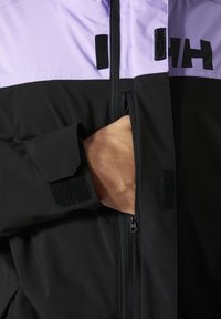Zwarte en lavendelkleurige jas met een ritsluiting, klittenbandaccenten en een hand in een zijzak. Gladde textuur en aansluitend ontwerp.