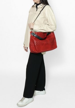 Sac à main tissé rouge avec une finition texturée, poignées en cuir noir et un design spacieux, porté avec une chemise beige et un pantalon noir.