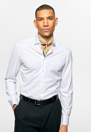 Mann med kort hår og øredobber har på seg en hvit knappet skjorte, beige halslue, svarte bukser og svart belte, står med en hånd i lommen.