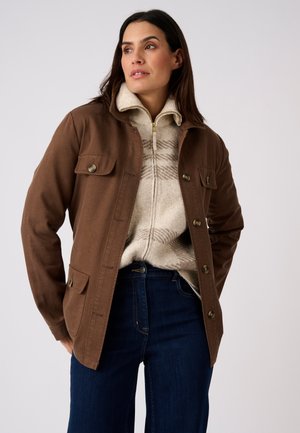Mujer con chaqueta marrón abotonada sobre un suéter beige con cremallera y estampado, y vaqueros azul oscuro, de pie con las manos en los bolsillos.