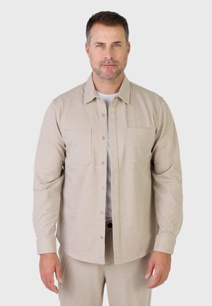 Mann med kort brunt hår og blå øyne, iført en beige knappet skjorte over en hvit T-skjorte, stående foran en ensfarget bakgrunn.