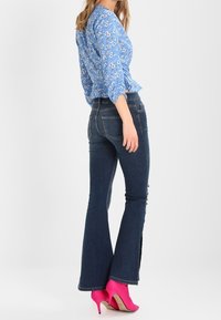 Blusa floreale blu con maniche a sbuffo, abbinata a jeans svasati scuri. Scarpe con tacchi alti di colore rosa brillante. Vista posteriore su uno sfondo chiaro.