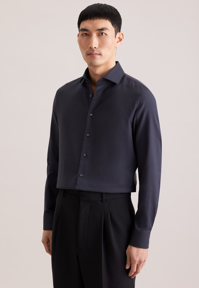 Homme portant une chemise ajustée en tissu texturé bleu marine foncé à boutons, manches longues, et un pantalon noir plissé, sur fond uni.